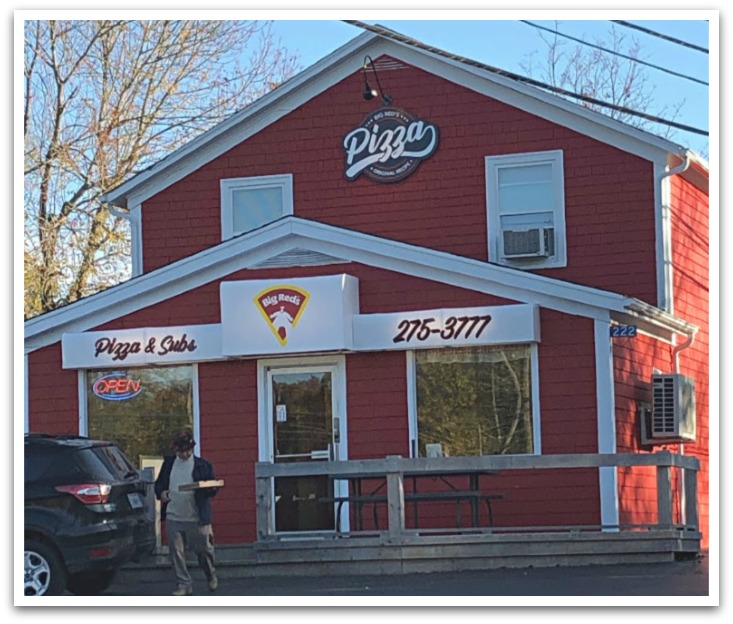 Man leaving Big Red's Pizza carrying a pizza box too his car. A red building with signs reading "Pizza & Subs", "Big Red's", and "275-3777". 