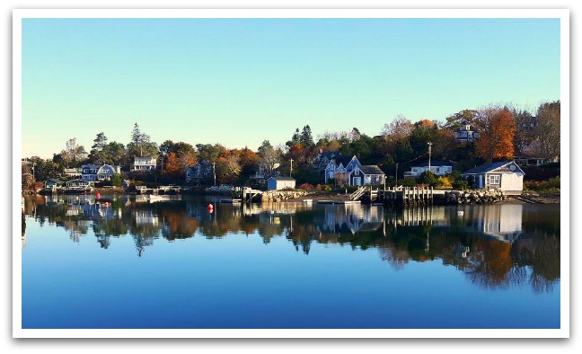 View of Water Street Chester showing wooden houses and wharves being reflected by the ocean in front of it with fall leaved trees in the background.