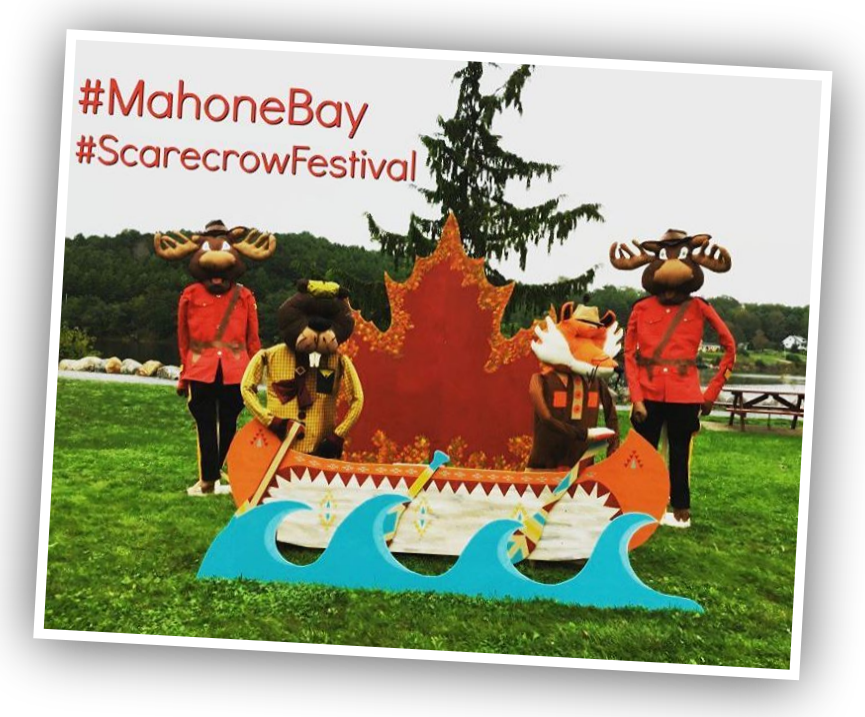 Life sized moose Mounty scarecrows behind a boat and sea prop with beaver scarecrows. Text reads "#MahoneBay. #ScarecrowFestival".