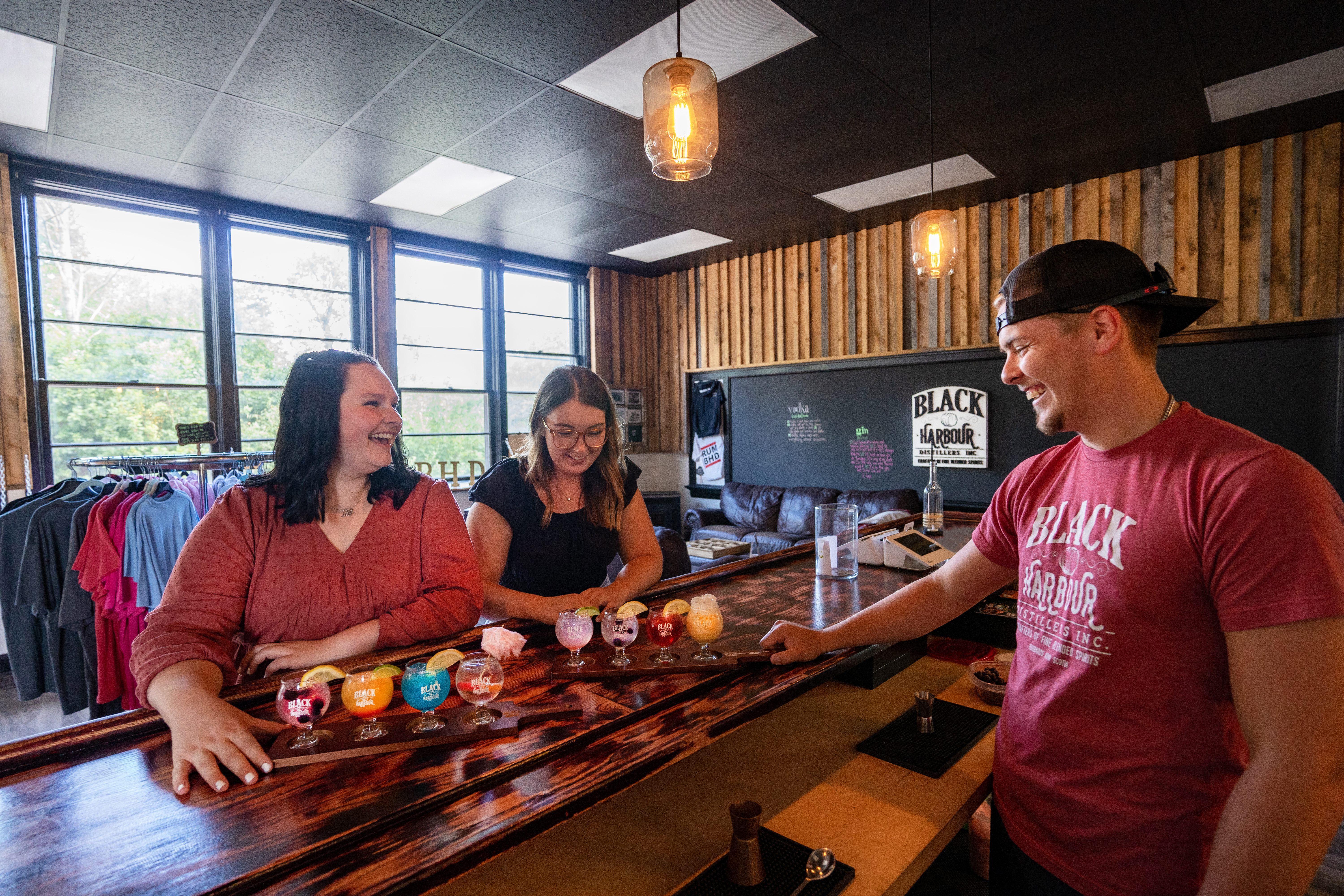 Man smiling passing two trays of sample colourful drinks to two smiling women at the Black Harbour Distillers bar..