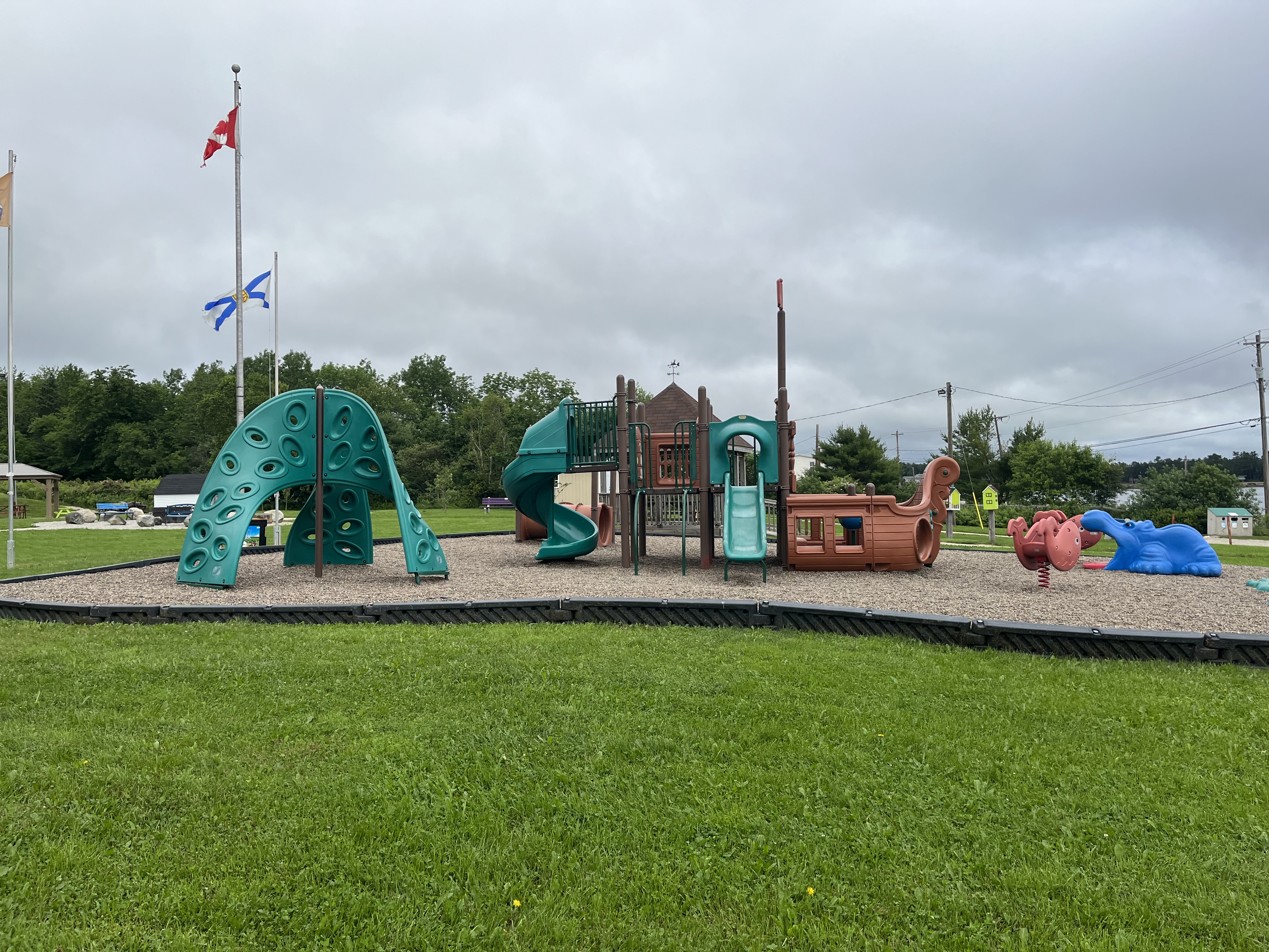 Pirate Ship Park with pirate themed play equipment on sand surrounded by grass. Canada and Nova Scotia flags on flag poles.
