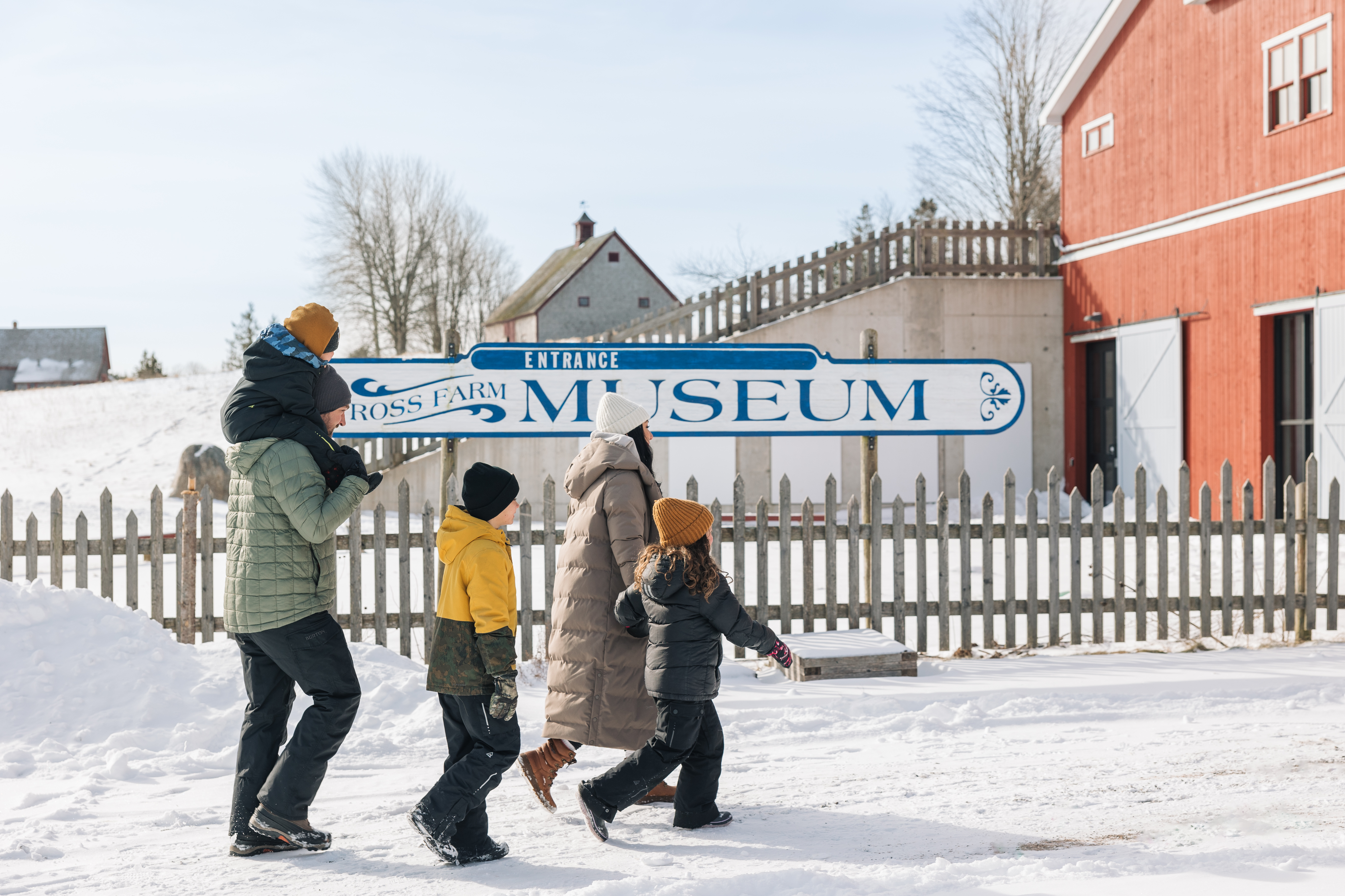 Family of five walking through the snow towards the entrance to Ross Farm with a sign reading "Ross Farm Museum" and a fence behind them.