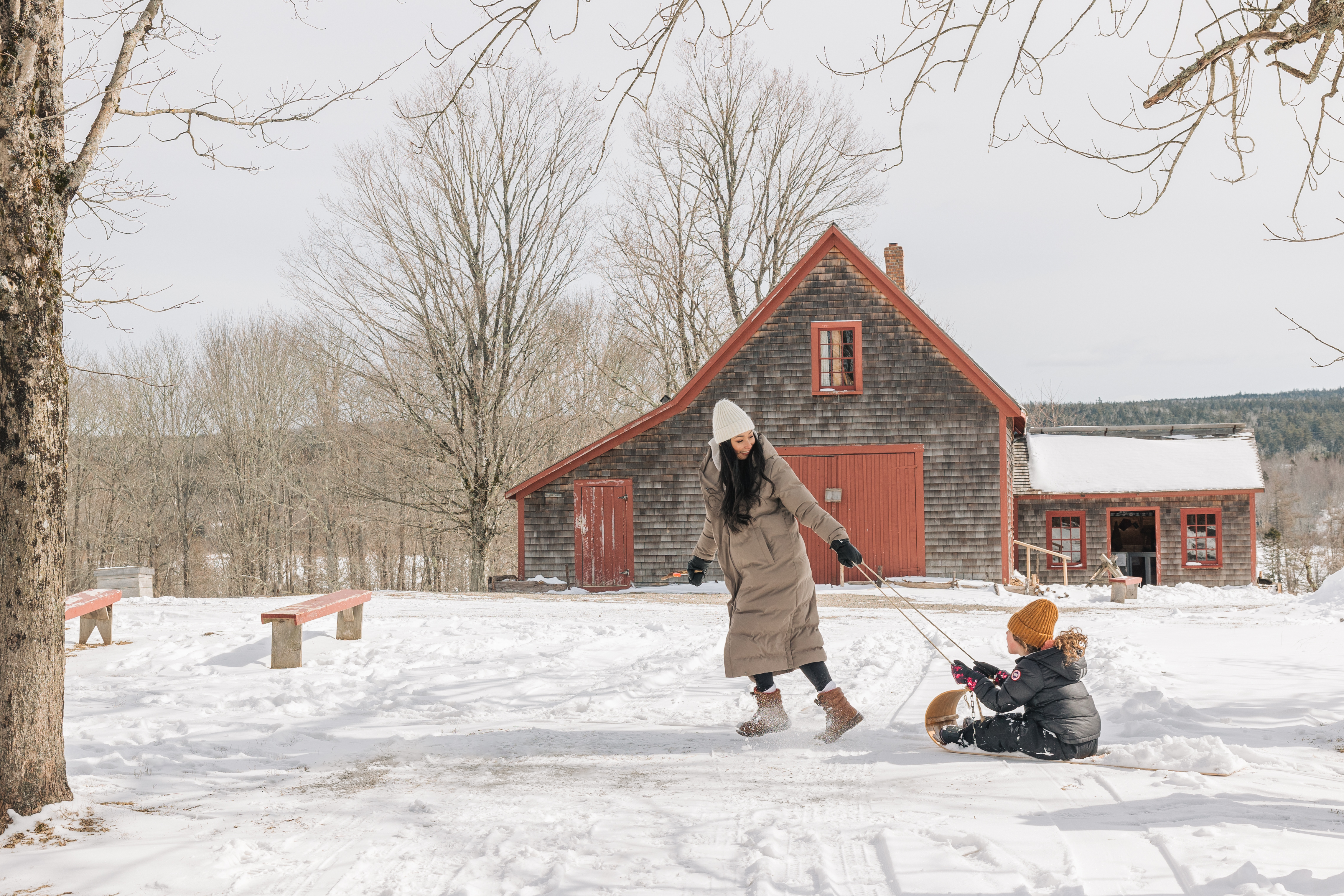 Woman pulling a child on a sled through snow with a brown and red barn in the background.