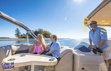 Couple sitting on a couch in the corner of a boat with the captain wearing a blue top steering the boat under blue sky with the sun shining and a small island in the background.