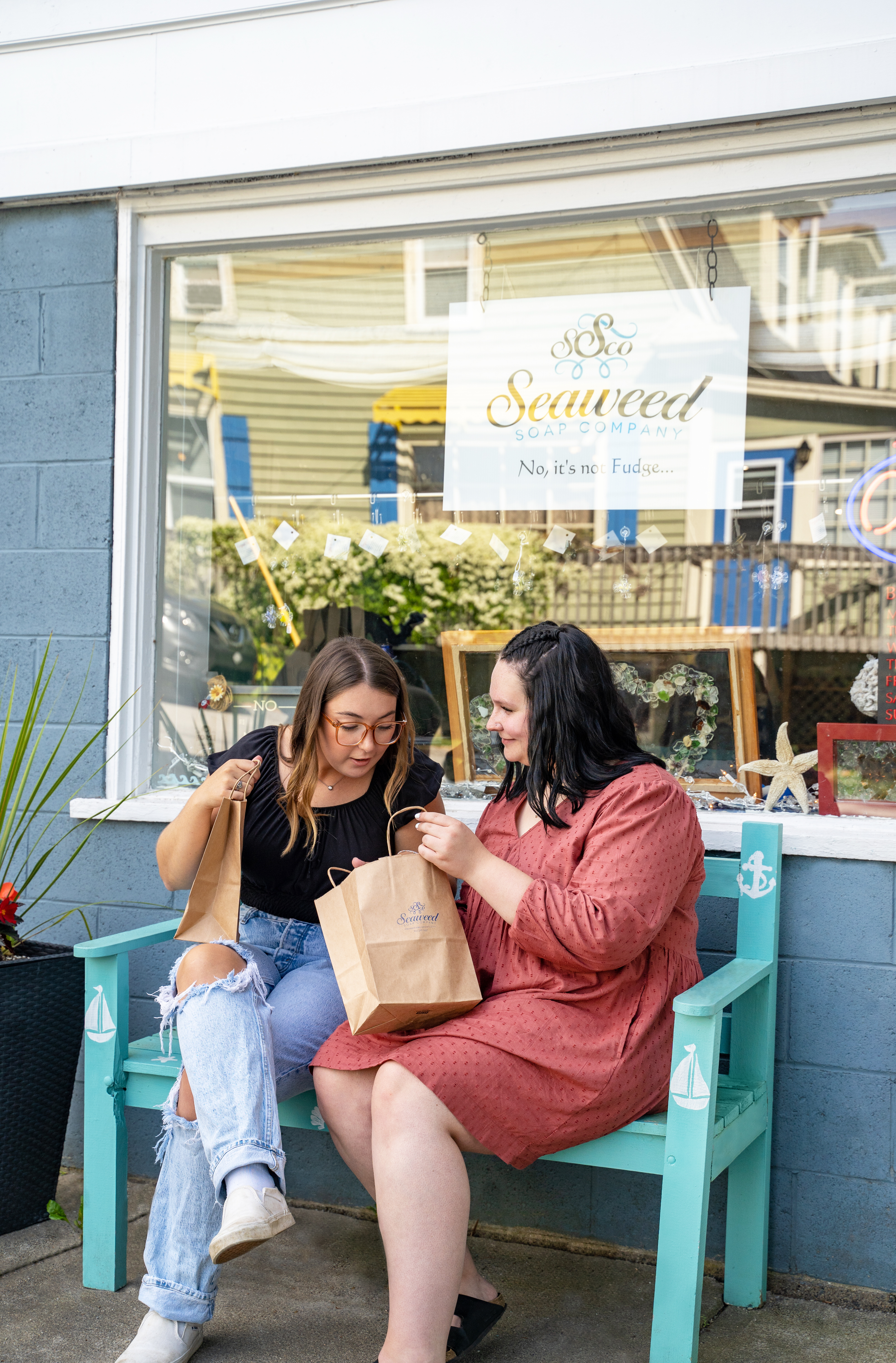 Two women sitting on an aqua bench outside of Seaweed Soaps shop looking at each other's purchases.