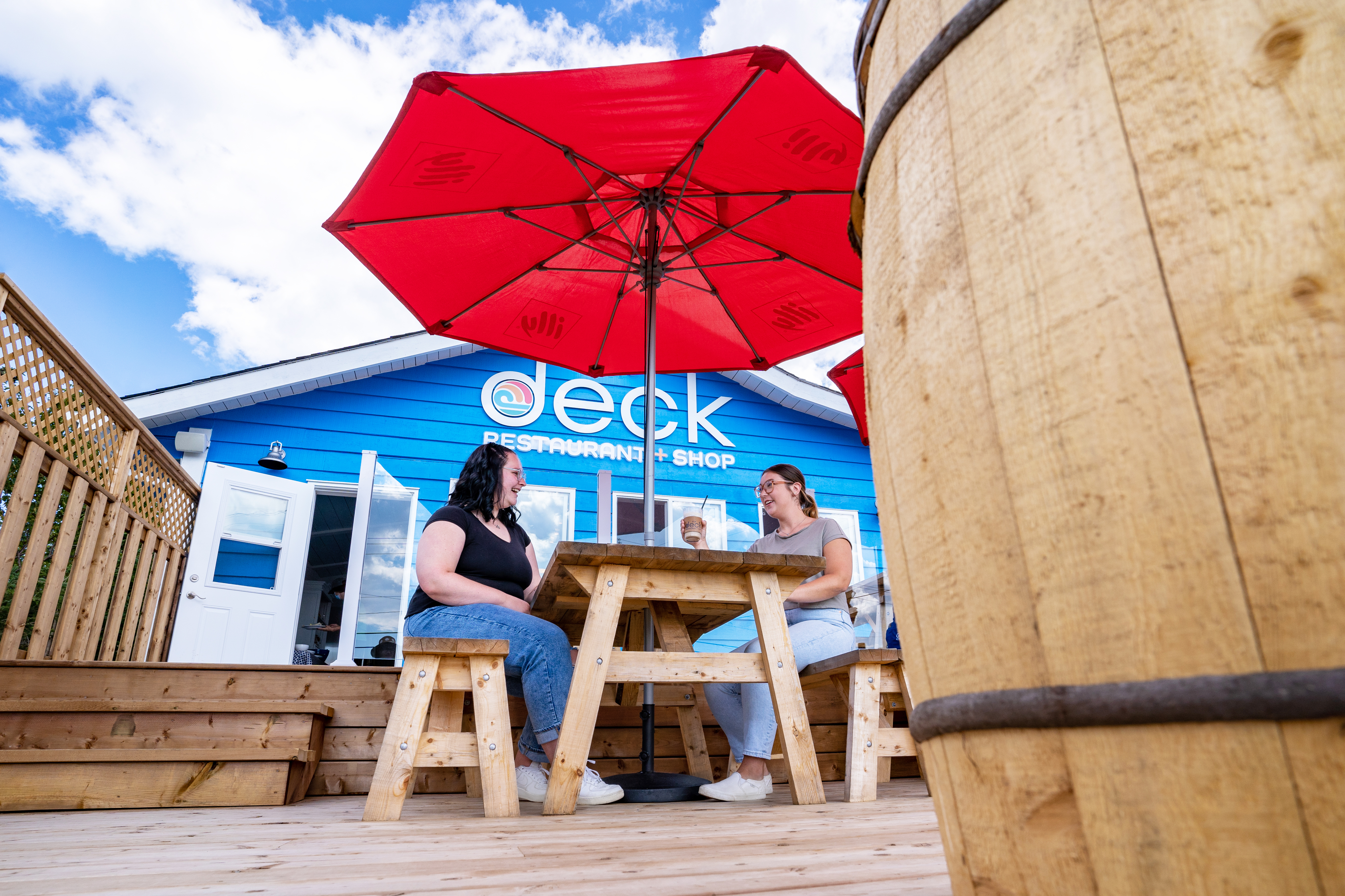 Two women sitting on a picnic bench under a red umbrella outside of The Deck - a blue building.