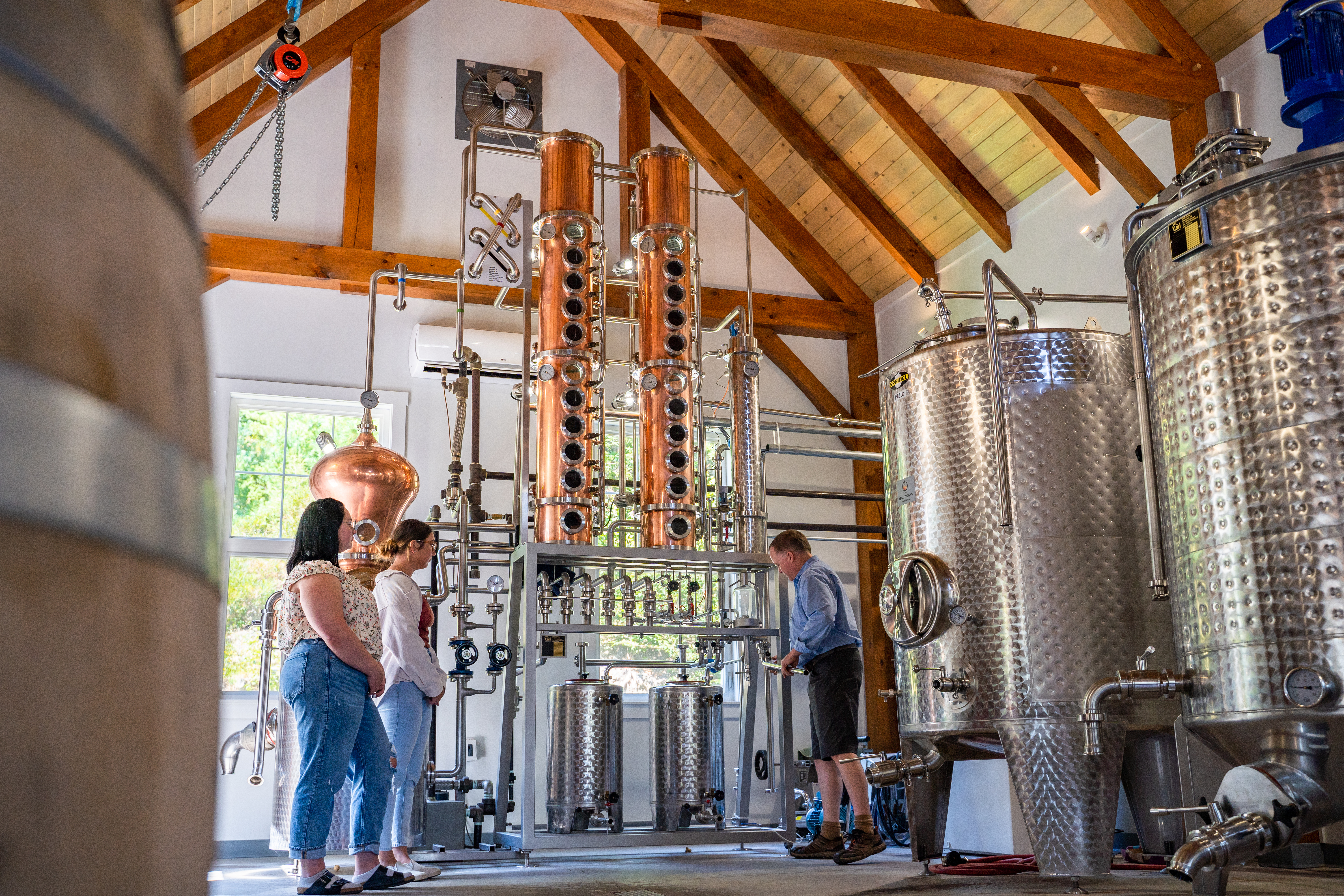 Tall metal distillers in a white vaulted room with a wooden ceiling. Man in a blue shirt giving a tour to two women.
