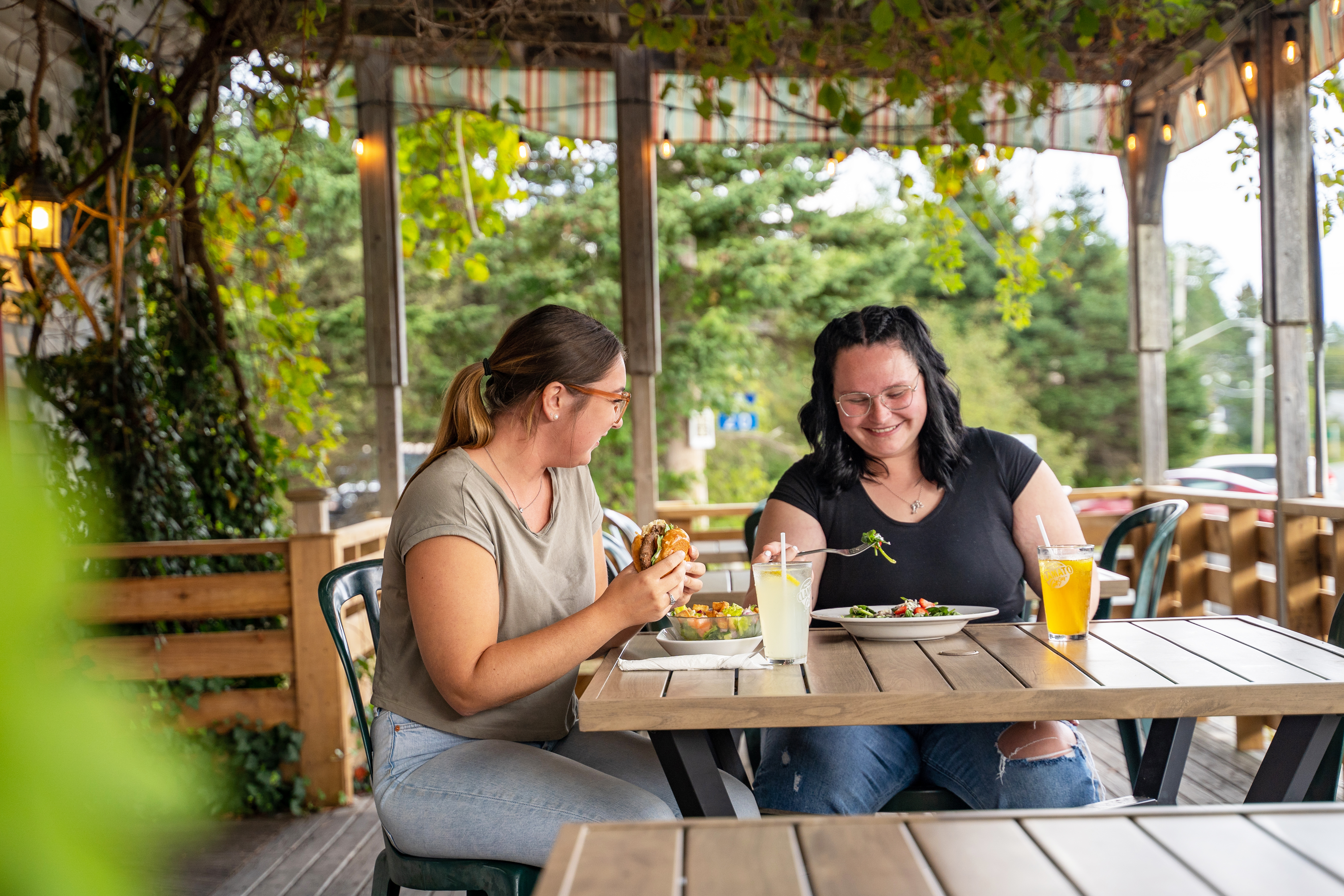 Two women sitting at a wooden table on The Trellis Cafe's porch with drinks and food. Ivy and fairy lights decorate the ceiling.