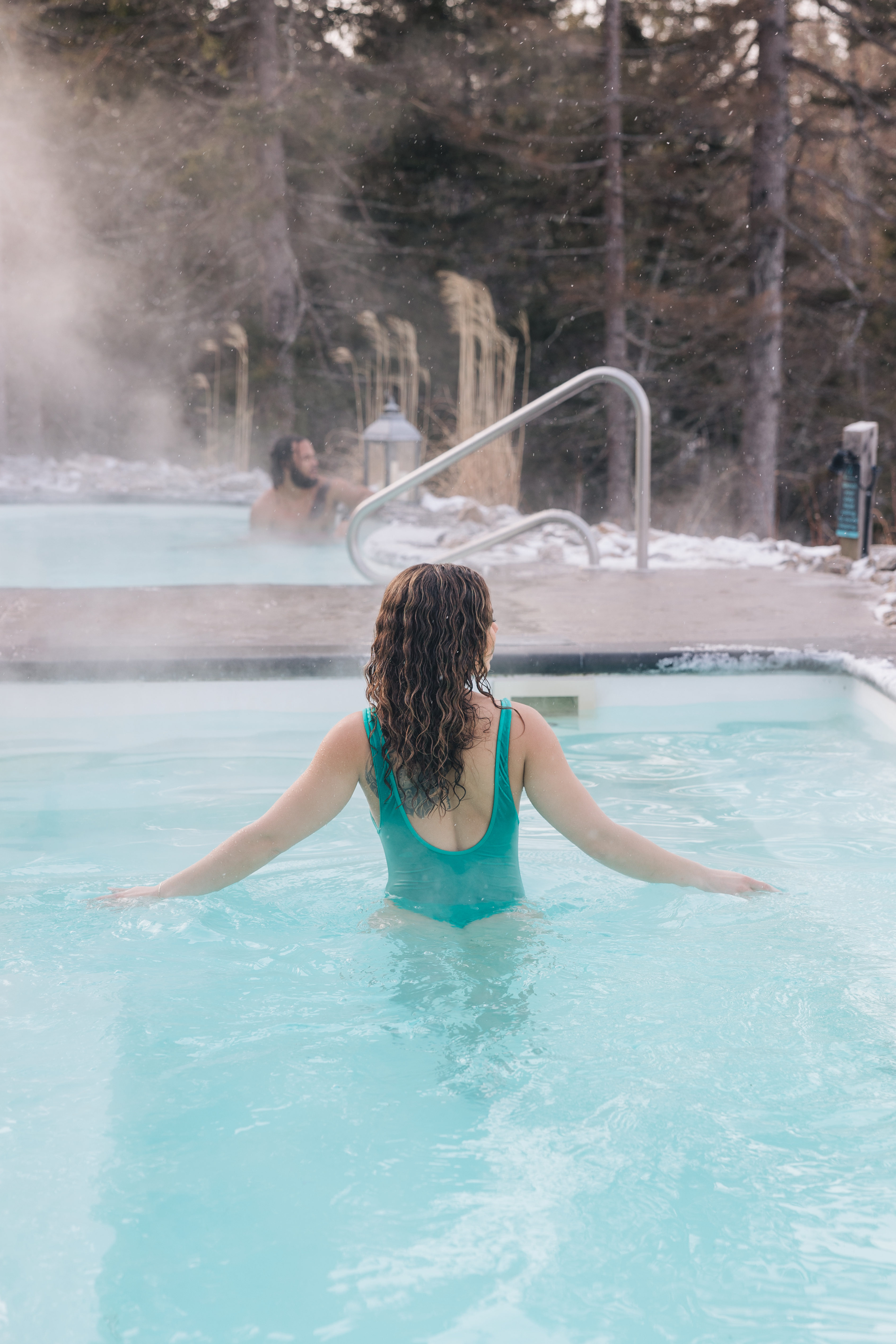 Woman in a hot tub with her arms out in winter wearing a green bathing suit. Man in a second hot tub in the background.