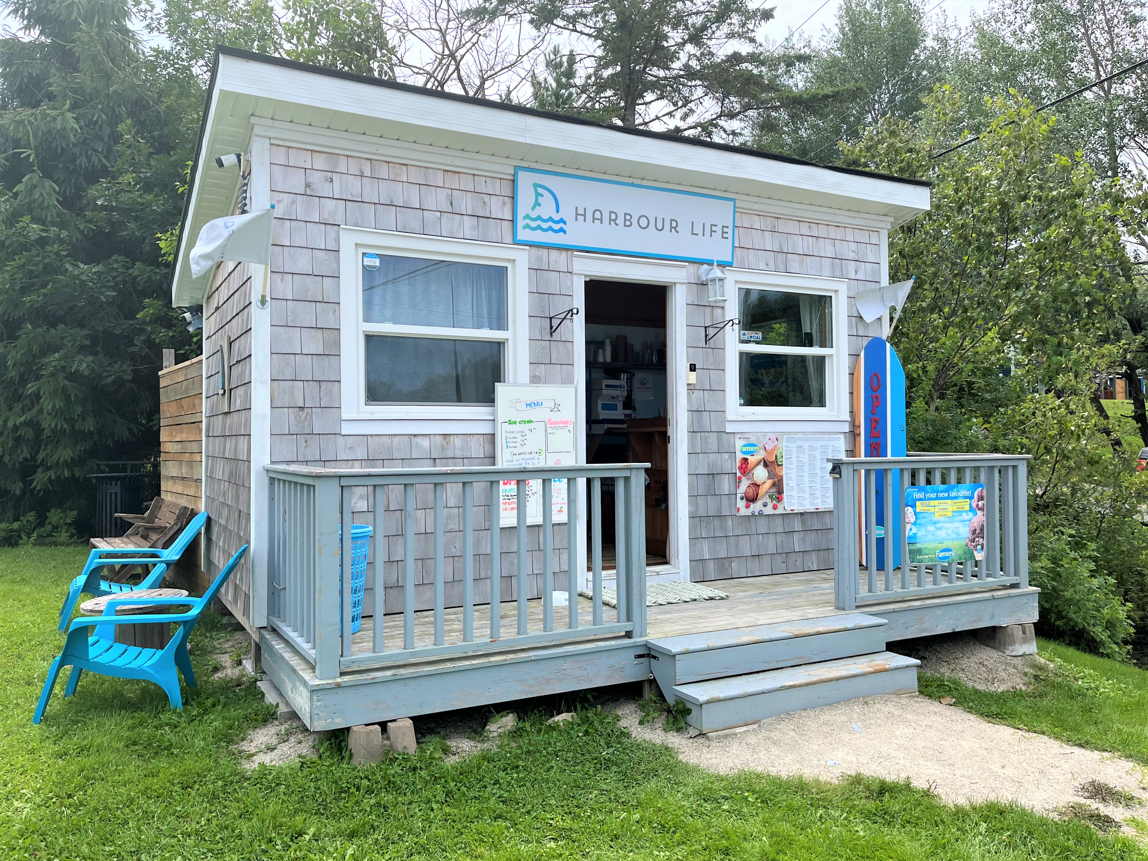 Small grey gabled building with a sign reading "Harbour Life" above the open door. A menu is outside showing the flavours of ice cream and two blue deck chairs are on the grass to the left of the building.