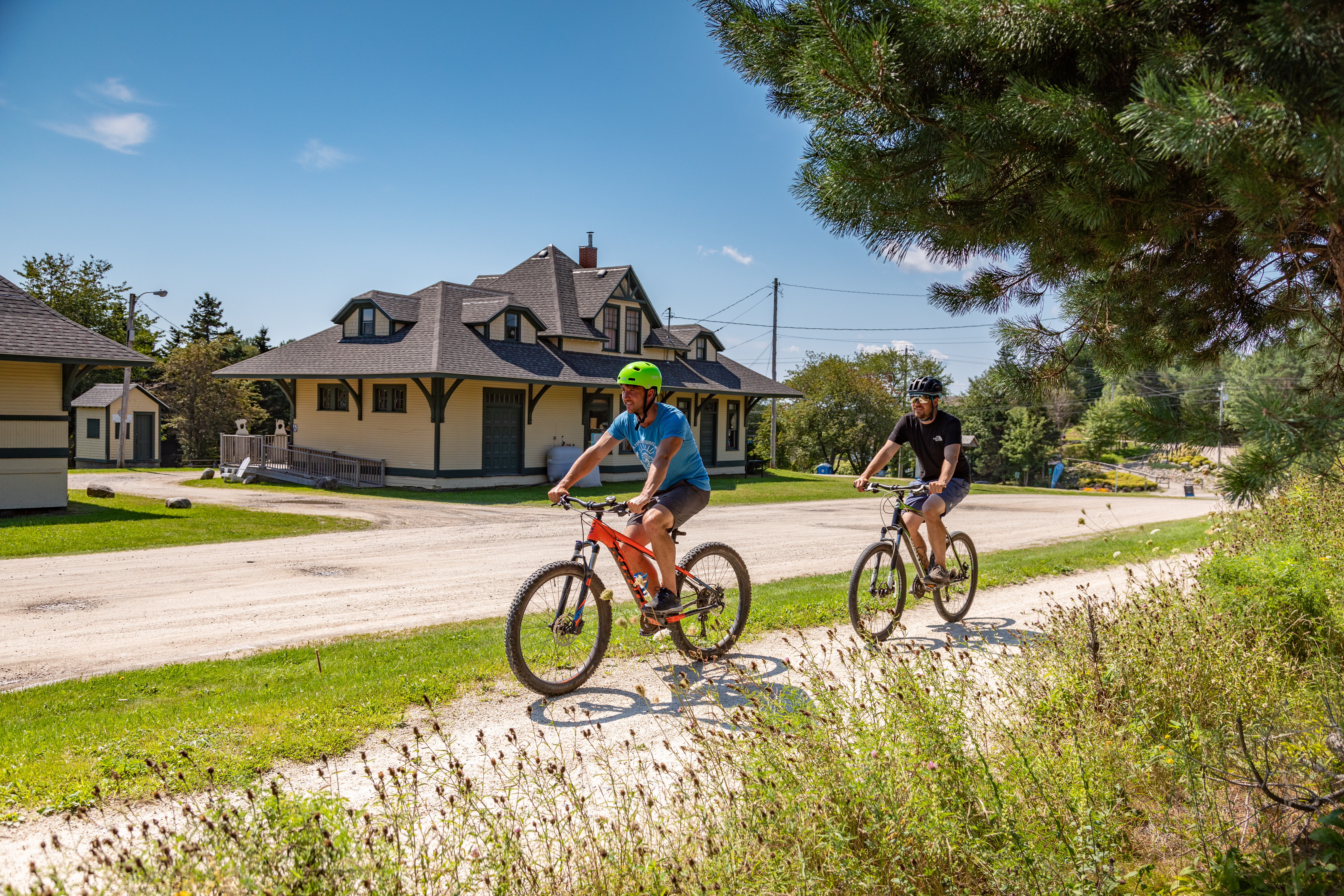Two people cycling past the Old Train Station along a gravel trail on a sunny day