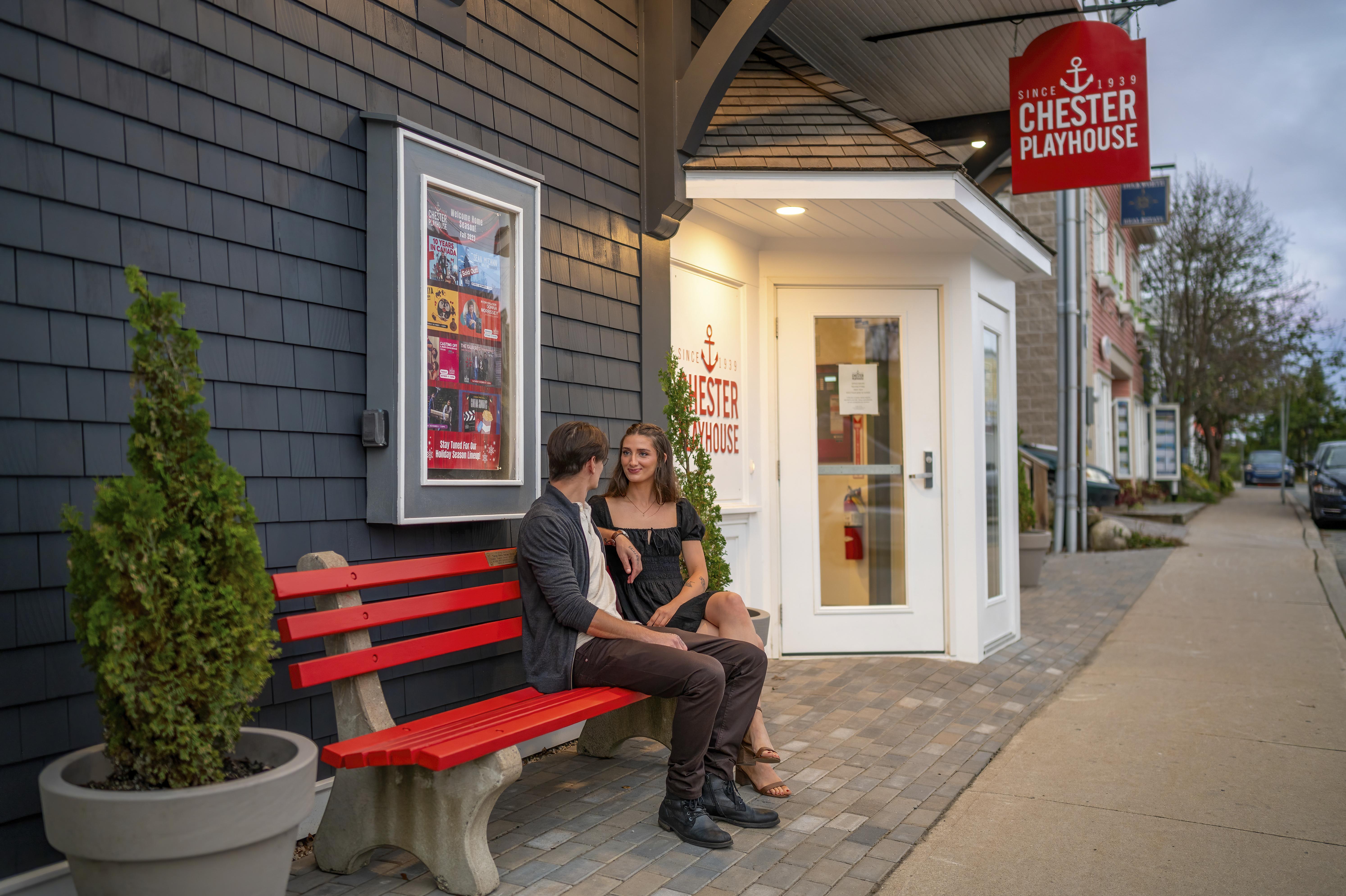 Two people sitting on a read bench outside the Chester Playhouse along the sidewalk.
