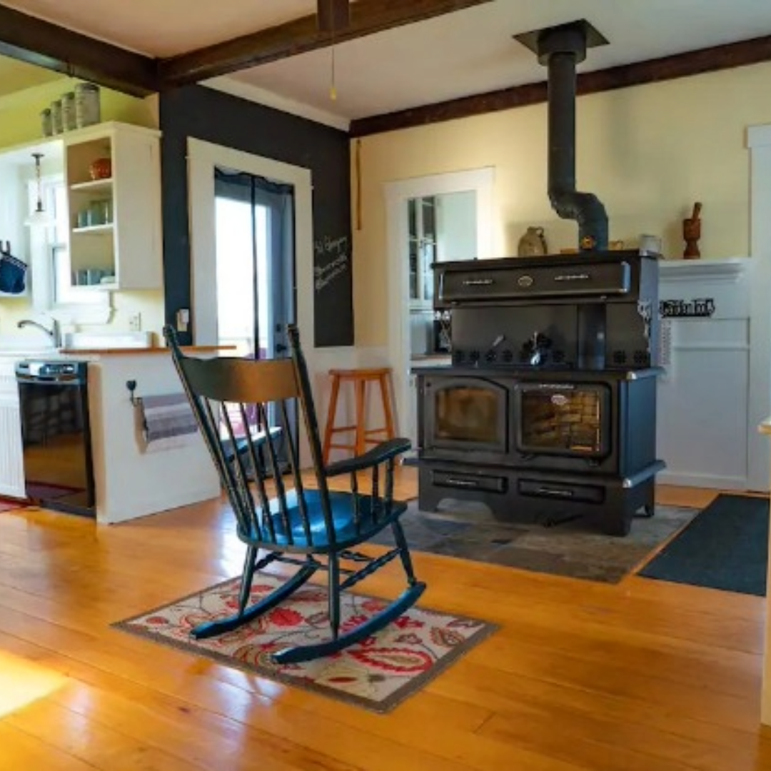 Photo of a rocking chair on warm wooden floors in front of a wood stove. Kitchen to the left.