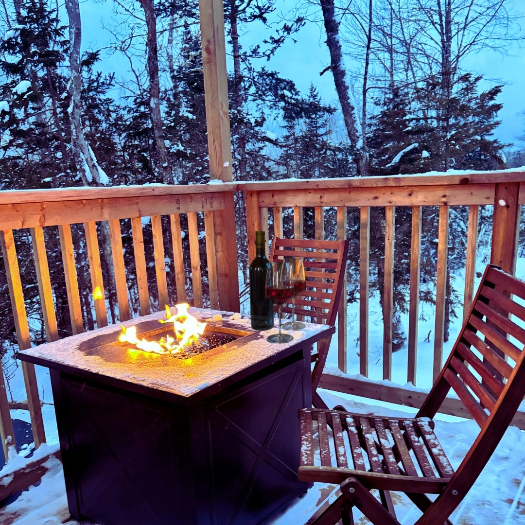 Photo of a wooden folding chair on a deck next to a propane firepit with a glass of wine surrounded by snow and trees.
