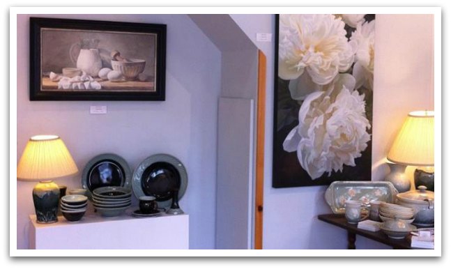 Amicus Gallery interior. Lamps on tables with pottery crockery and a painting of baking utensils on the mall.