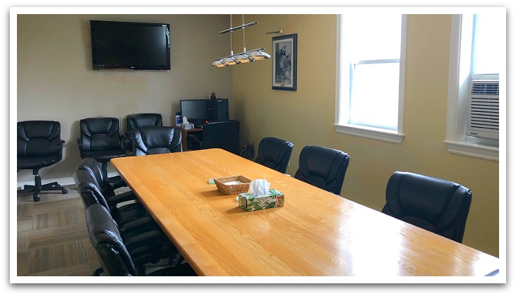 A small room with a rectangular wooden table by two windows on a carpet. Black office chairs surround the table with extras underneath a wall mounted TV.