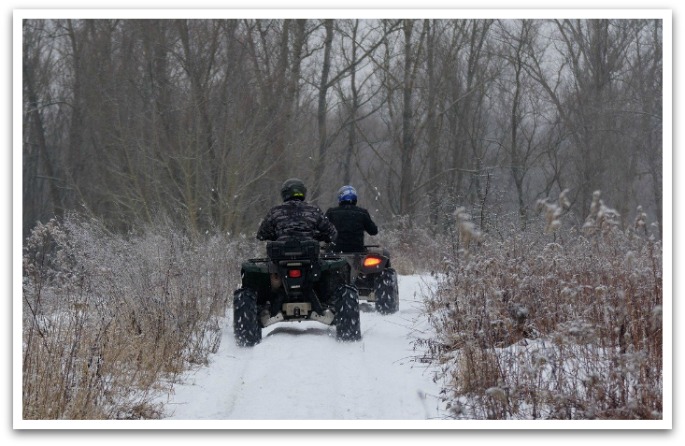 Two men riding TAVs down a snow covered path with trees in the background.