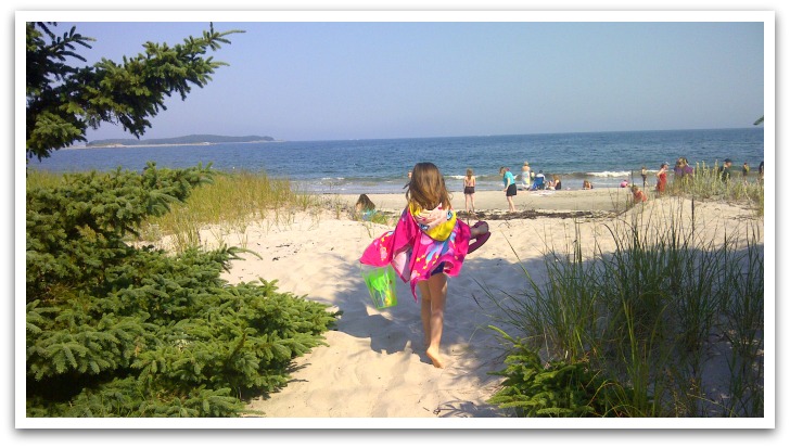 Girl wearing a pink hooded towel carrying a bright green bag walking on sand towards the ocean.