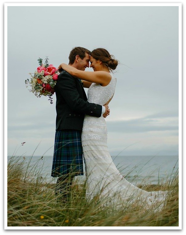 Bride and groom embracing. Bride wearing a white wedding dress holding a pink bouquet, the groom is wearing a kilt. Long grass and ocean in the background.