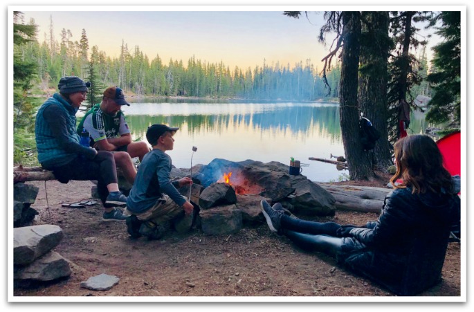 Family sitting around an open fire roasting marshmallows next to a tent by a lake