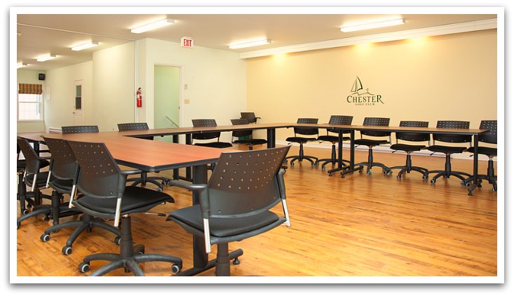 A cream painted well lit room with wooden floors. Tables are in a U shape with 14 places.