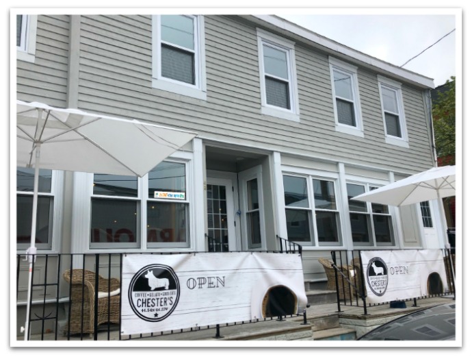 Exterior of the Jib with flags on the patio barriers reading "Chester's, open". Patio furniture is on the patio in front of the beige building.