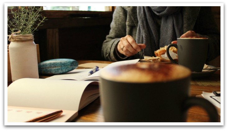Person with a croissant and cup of coffee wearing a scarf sitting at a table in a cafe. Paper and pens and a plant are on the table.