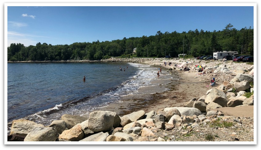 Rock lined sandy Fox Point Beach with groups of [people sitting under umbrellas. Houses and trees in the background.