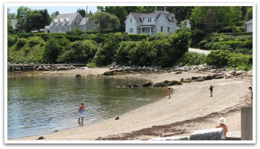 Small sandy beach with small children and a lady lined with rocks following a small road leading to white houses.