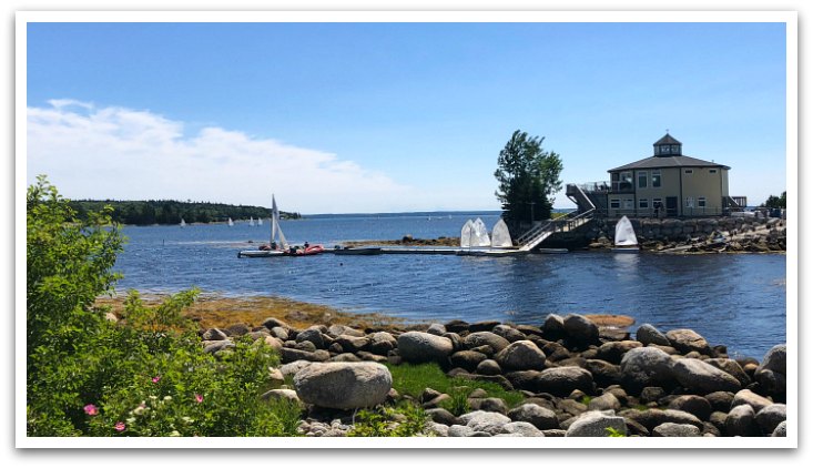 Yellow hexagonal Hubbards Sailing Club building with sailing boats attached to the wharf outside and more in the bay. Rocky beach and trees are in the foreground.