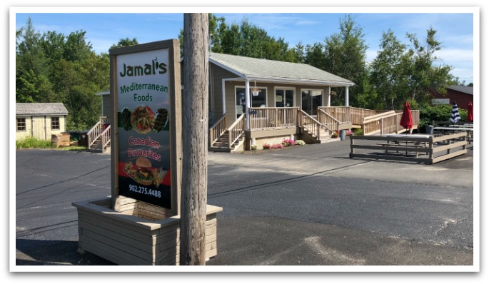 Sign reading "Jamal's Mediterranean Foods. Canadian Favourites. 9022754488." Next to a beige building with parking, patio furniture, and umbrellas.