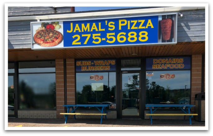 Exterior of a strip mall shop. A blue sign on the building with photos of a pizza and donair meat skewer reads "Jamal's  Pizza 275-5688". Windows of shop have text reading "Subs, Wraps, Burgers, Donairs, Seafood". Two blue benches are outside.
