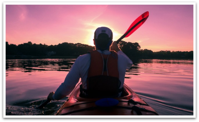 Man wearing a white hat and red life vest paddling in a lake at sunset.