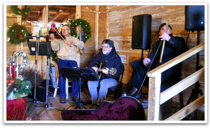 Group of three old men playing string instruments on a small stage decorated with Christmas wreaths and lights in the Hubbards Barn. 
