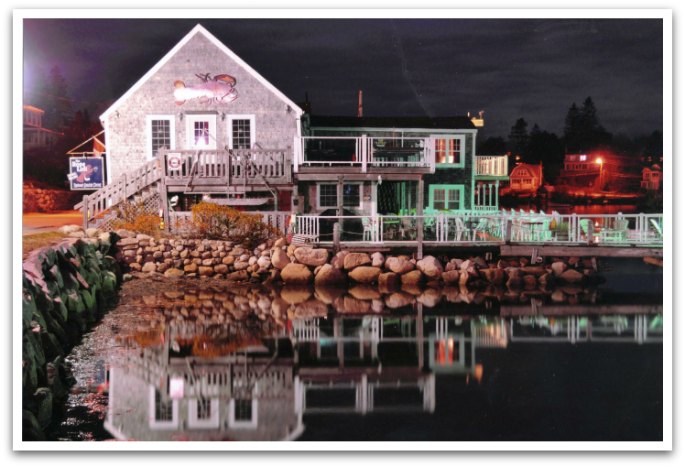 The Rope Loft pub at night, a two story grey building with a large deck area by the bay reflecting in the water.