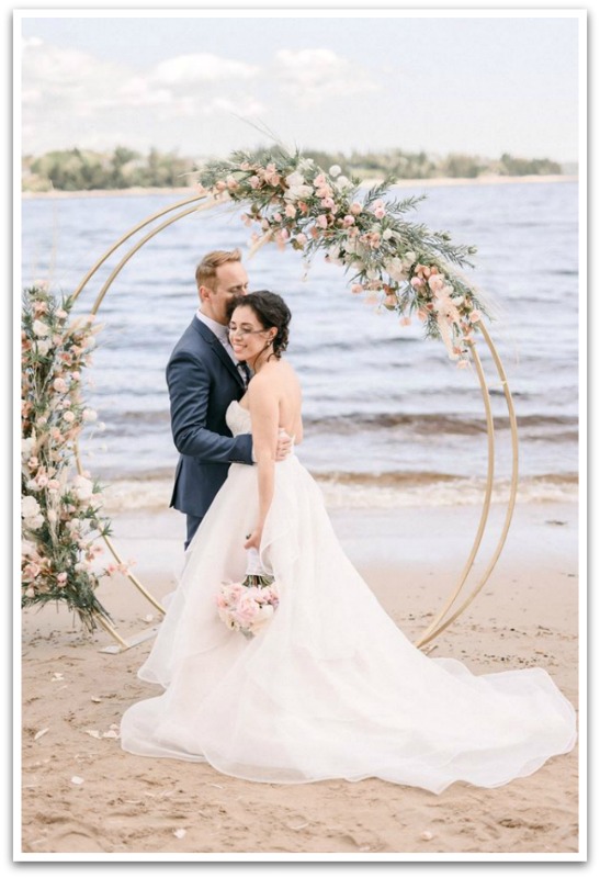 Bride and groom in front of a decorative circular prop with flowers attached on a beach.