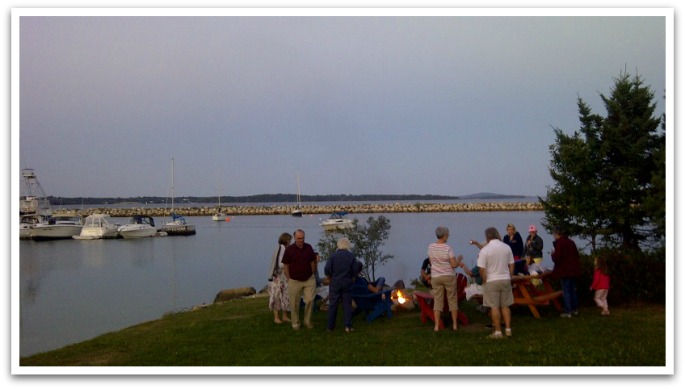 Group sitting around a bonfire by the ocean with boats in the background