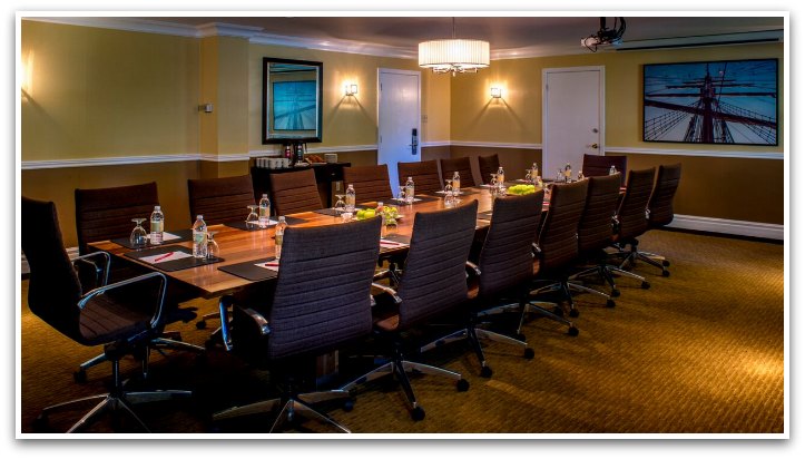 Oak Island Resort Boardroom with yellow and burgundy wallls, a brown carpet, a long wooden table with water bottles by each seat, 16 brown office chairs.