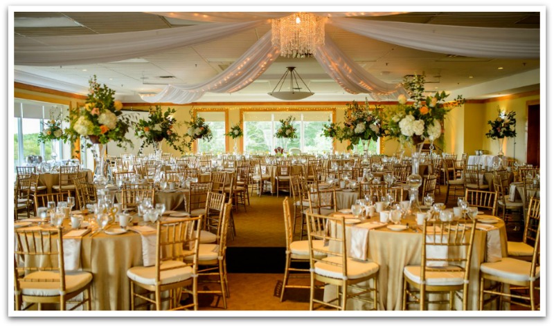 Ding hall with round tables and cushioned dining chairs with flower boquests and decorative white canopies surrounding the chandelier. Yellow walls with many windows.