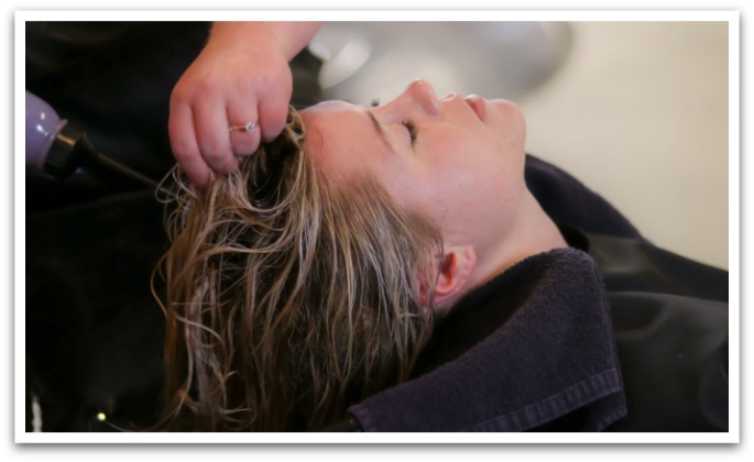 A woman lying back in a hair salon sink with a black towel around her shoulders with someone pouring toner out of a purple bottle on the client's  hair.
