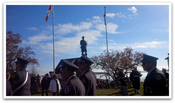 Men in military uniform marching past a statue of a highland soldier next to a Canadian and Nova Scotian flag.