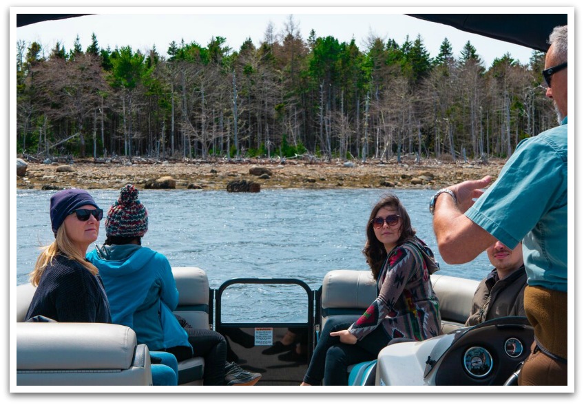 Man speaking to a group of 4 on a boat wearing hats with the shore and trees in the background.