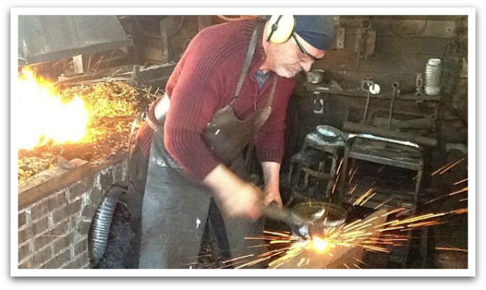 Man blacksmithing with sparks coming from metal with a brick oven behind him.