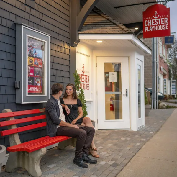 Photo of two people sitting outside the Chester Playhouse on a red bench, entrance in the background