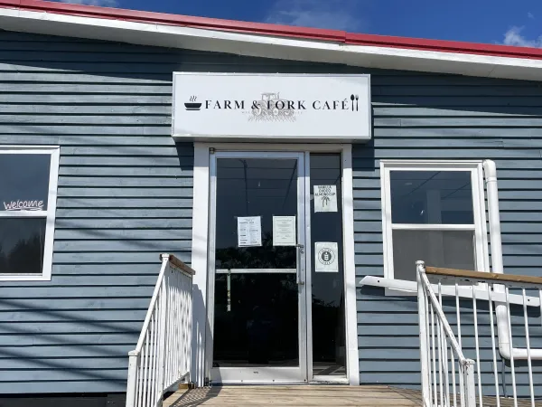 Photo of the entrance to Farm & Fork Cafe showing a wooden steps, wooden ramp, while railing, glass door, blue building, and sign.