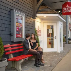 Photo of two people sitting outside the Chester Playhouse on a red bench, entrance in the background