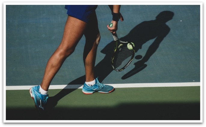 Woman wearing blue sneakers bouncing a tennis ball on a court to serve, photo taken from her hips down.