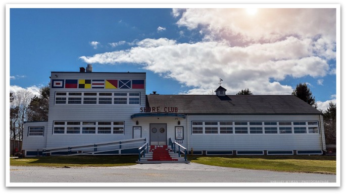 The Shore Club exterior. A white, large, two story building with sailing flags. A ramp and stairs lead to double doors with "Shore Club" letters above the doorway.