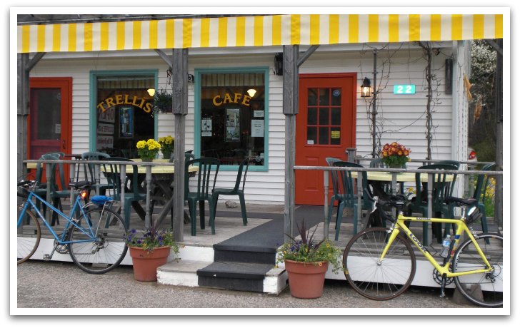 The Trellis Cafe Hubbards exterior. Shows white building with a red door and a yellow striped shade over the patio. Bikes are leaning against the rails.