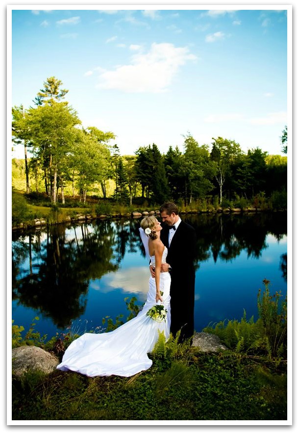 Bride and groom posing by a glass lake on a sunny day with trees and greenery behind them.